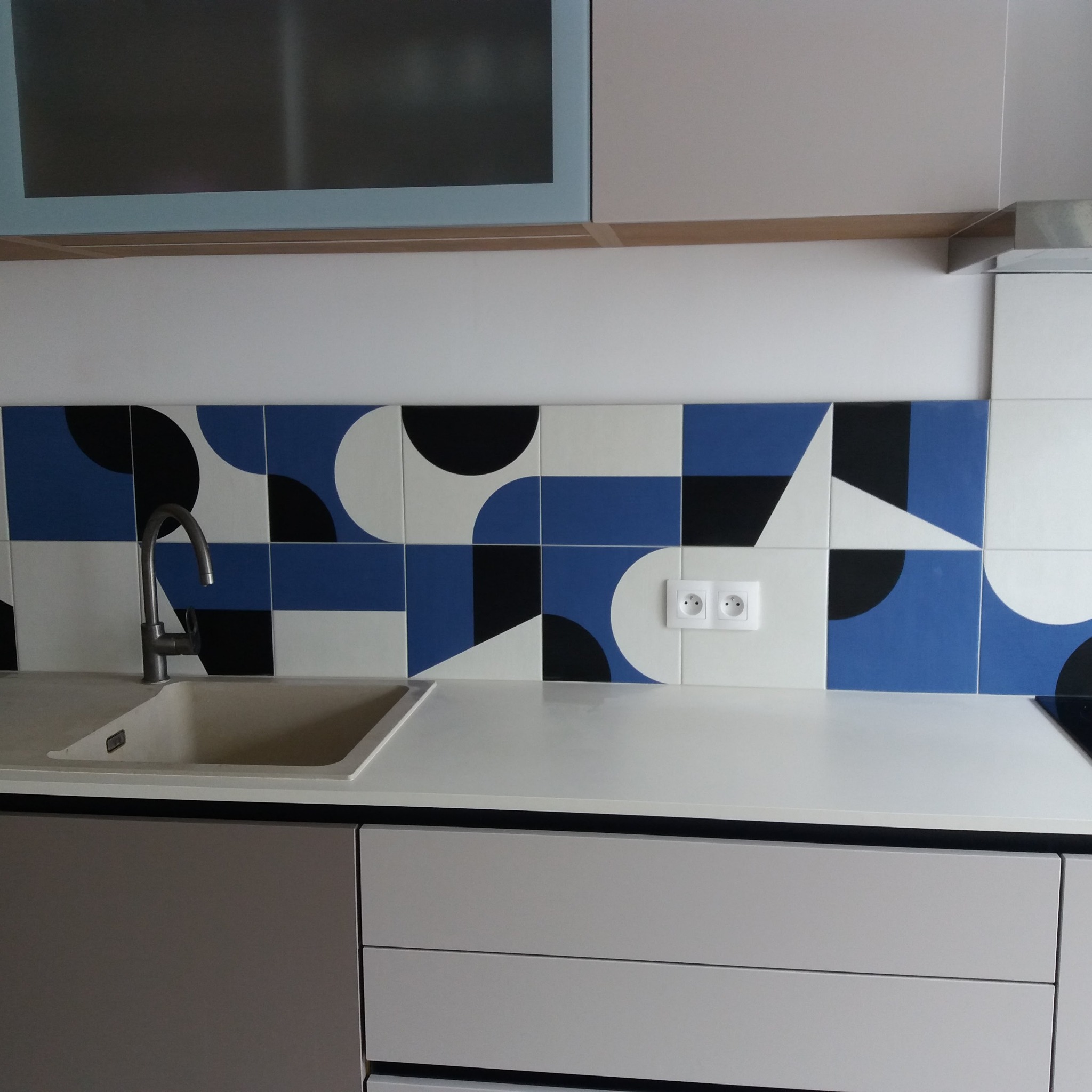 Fragment kuchni z nowoczesną zabudową: szare szafki dolne, biały blat, umywalka z szarego konglomeratu i dekoracyjne płytki na ścianie w kolorach białym, czarnym i niebieskim z geometrycznym wzorem.