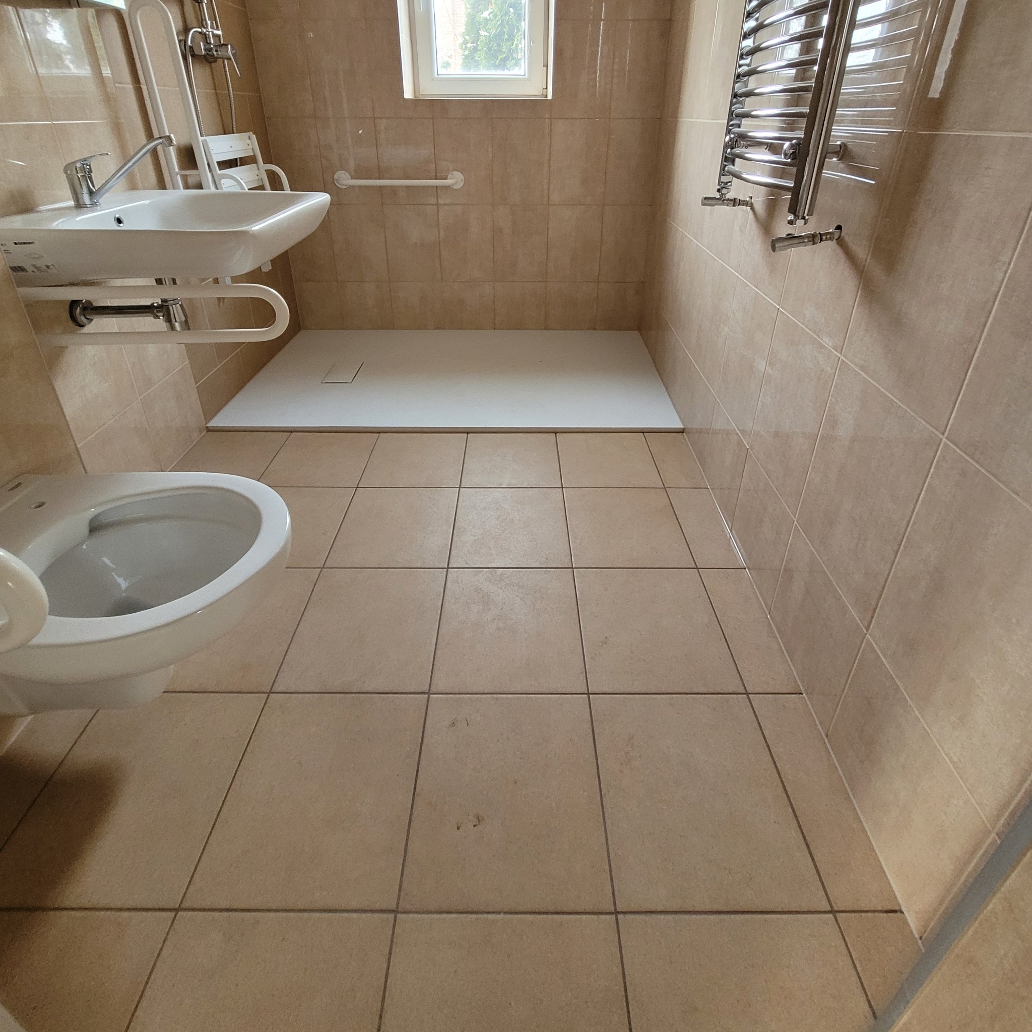 Beżowa łazienka z białą umywalką na regulowanej wysokości z poręczami, toaletą, brodzikiem bezprogowy i grzejnikiem drabinkowym