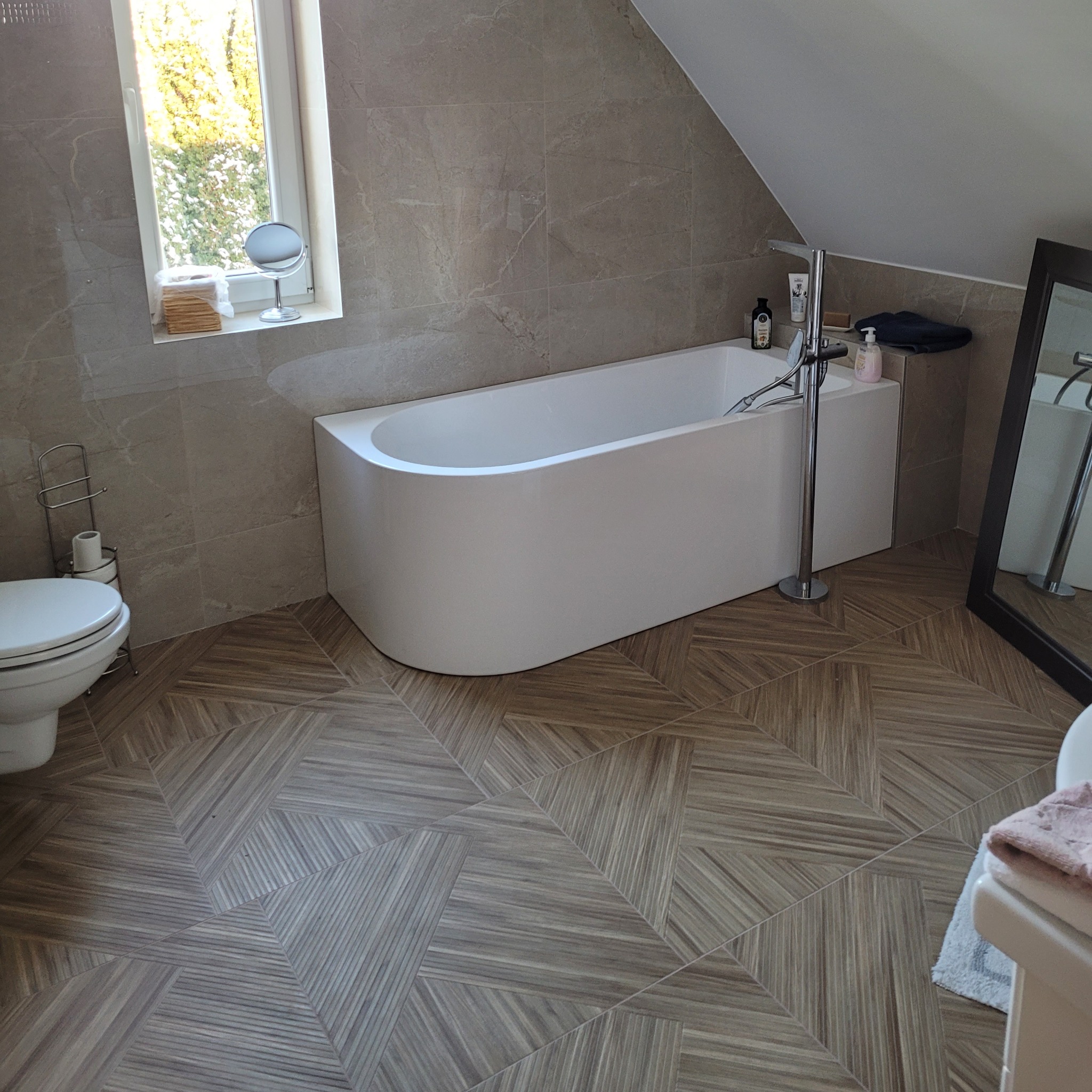 Nowoczesna łazienka na poddaszu z płytkami podłogowymi w geometryczny wzór, wolnostojąca wanna, umywalka i lustro. Okno z widokiem na zieleń.