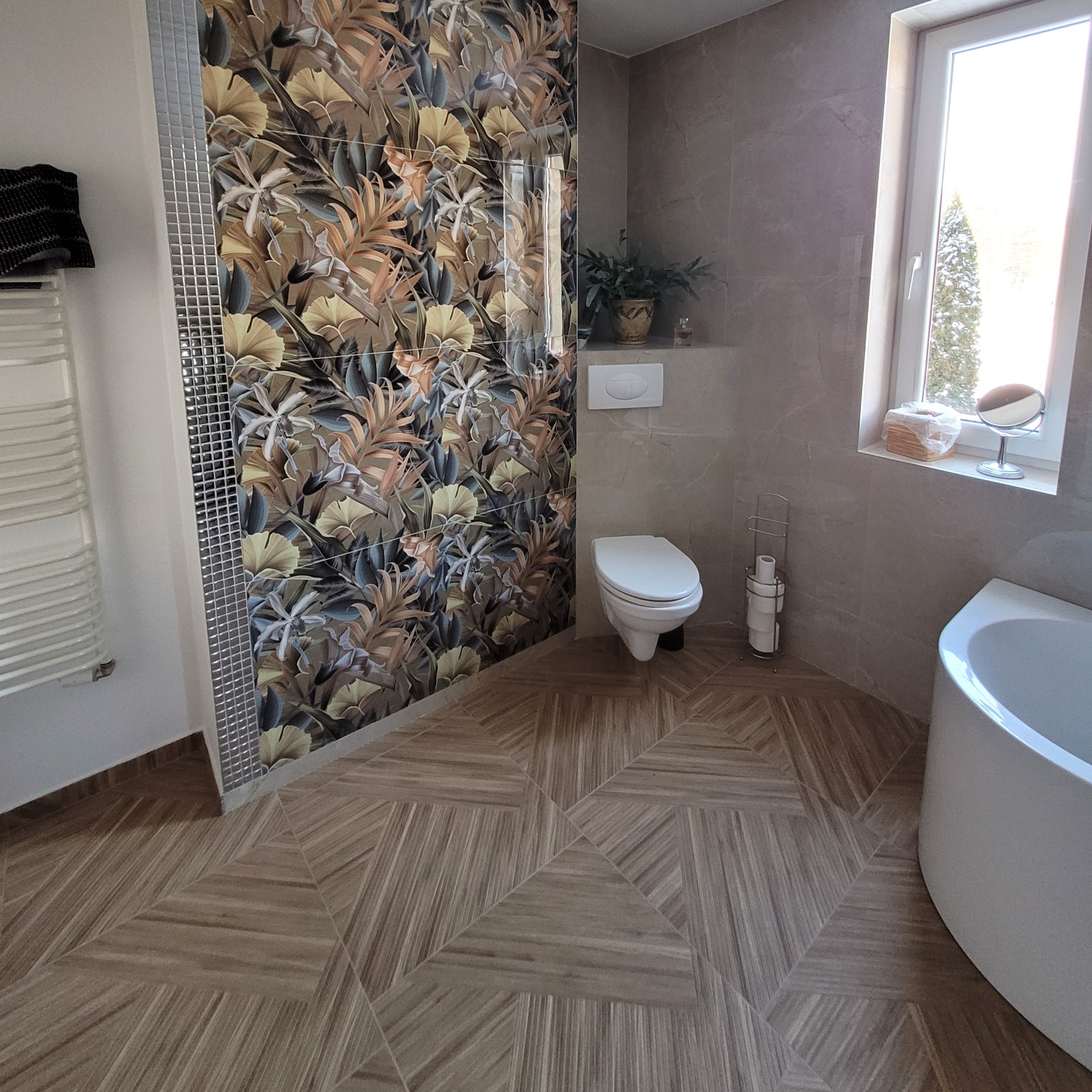 Wykończona łazienka z toaletą, wanną i grzejnikiem; ściana z wzorzystymi płytkami w stylu tropikalnym, podłoga z płytek ułożonych we wzór geometryczny, okno z widokiem na zaśnieżone drzewo.