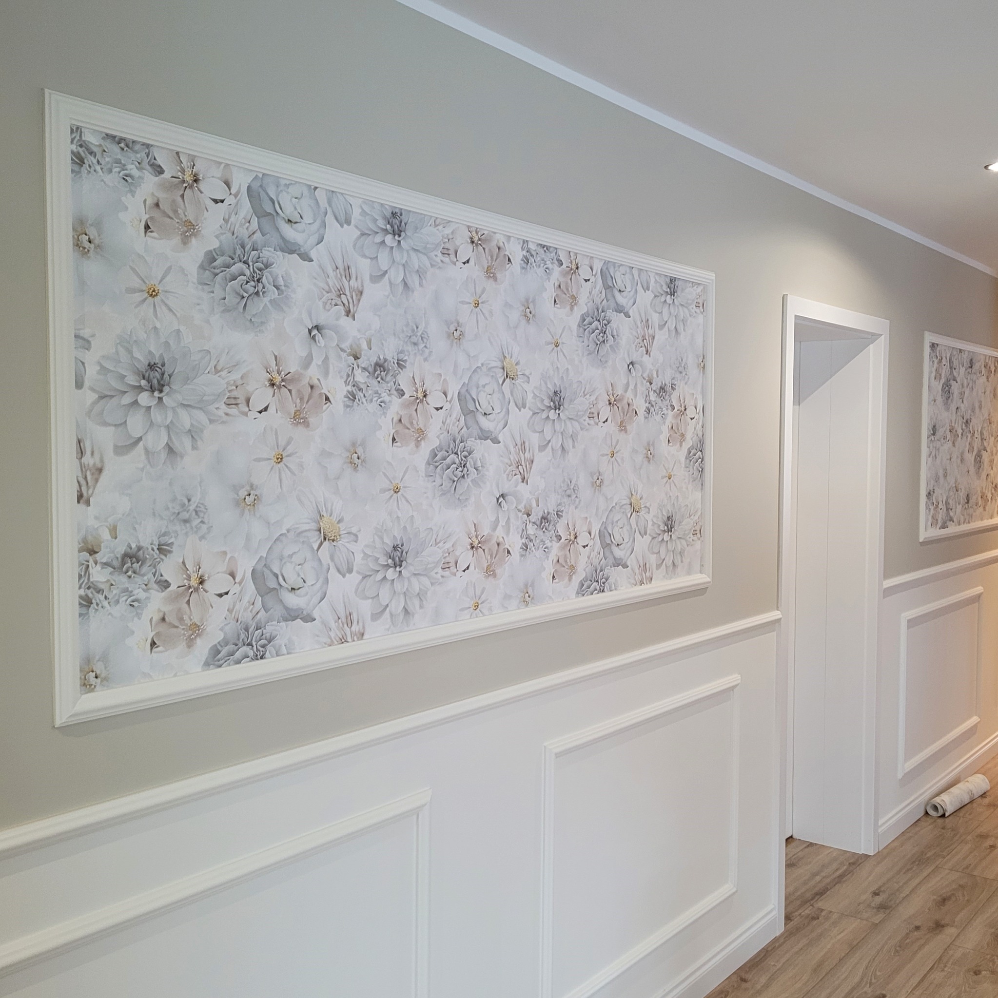 Elegancki korytarz z panelami ściennymi w stylu angielskim, ozdobiony kwiatową tapetą w ramie i drewnianą podłogą, z widoczną rolką tapety w rogu.