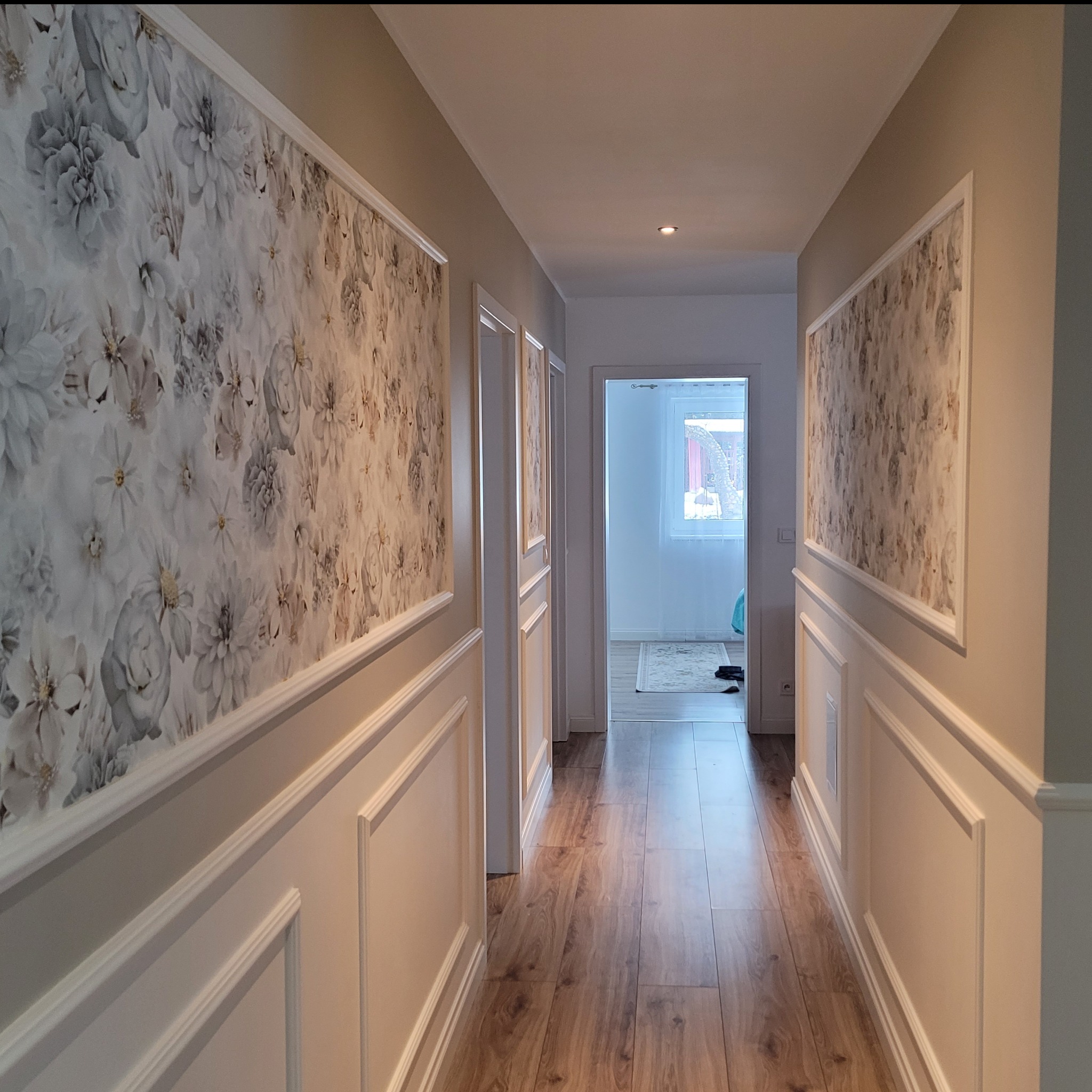 Korytarz po remoncie z panelami ściennymi z dekoracyjną tapetą w kwiaty i drewnianą podłogą, widok na otwarte drzwi do pokoju z dywanem i oknem.