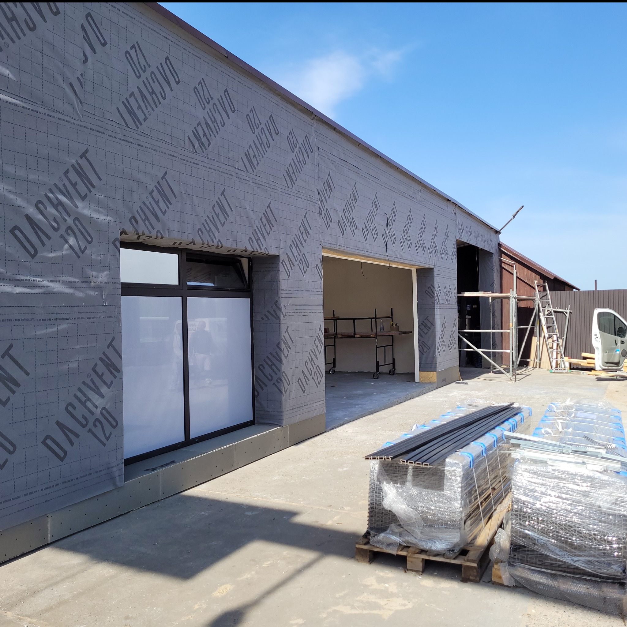 Budynek w trakcie ocieplania, widoczna szara membrana Dachvent 120 na ścianach, zamontowane okna z czarnymi ramami, w tle rusztowania i zaparkowany biały van, na pierwszym planie palety...