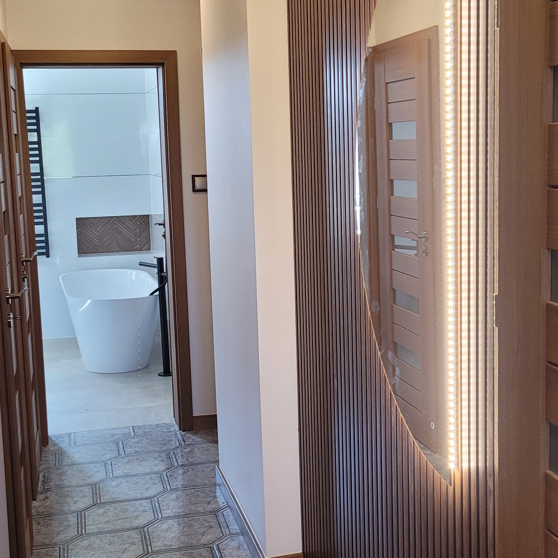 Nowoczesny korytarz z widokiem na łazienkę: biała wanna wolnostojąca, heksagonalna podłoga, drewniana okładzina ścienna i podświetlane lustro.