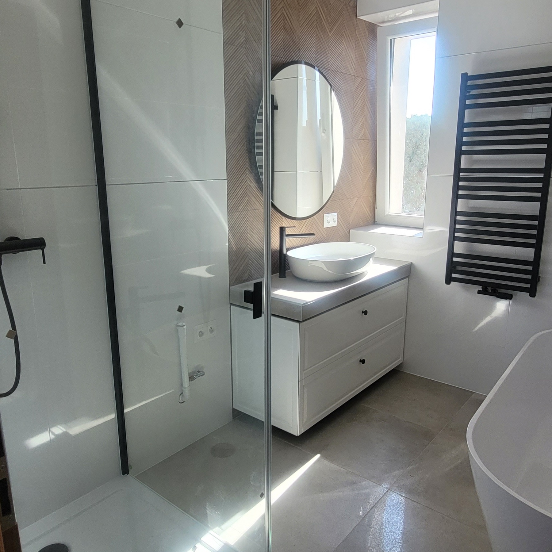 Nowoczesna łazienka z kabiną prysznicową za szklaną ścianką, owalnym lustrem, białą szafką z umywalką nablatową i grzejnikiem drabinkowym w czarnym kolorze.