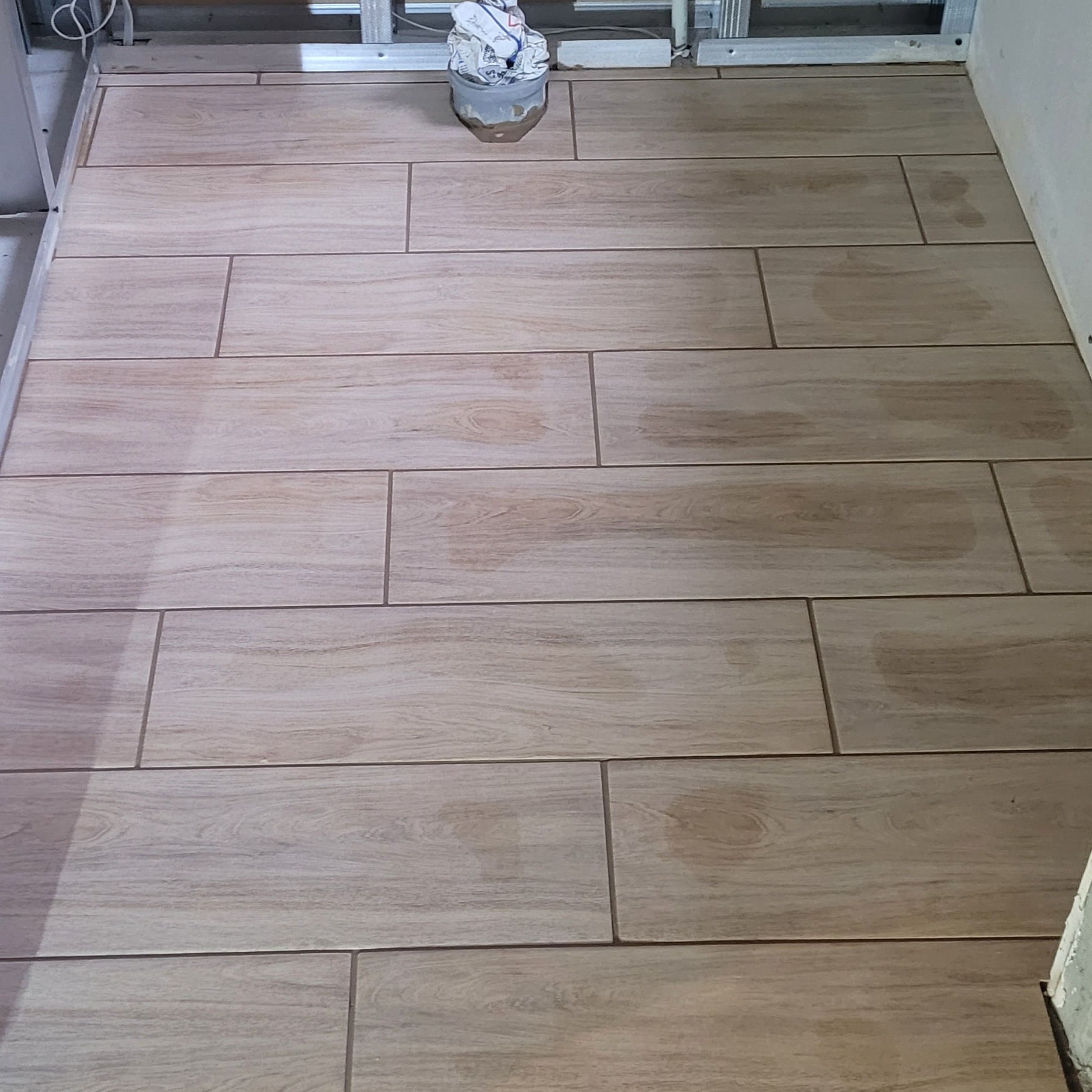 Ułożone, podłużne płytki podłogowe imitujące drewno w trakcie prac wykończeniowych w łazience, widoczny metalowy stelaż i wiadro z zaprawą.