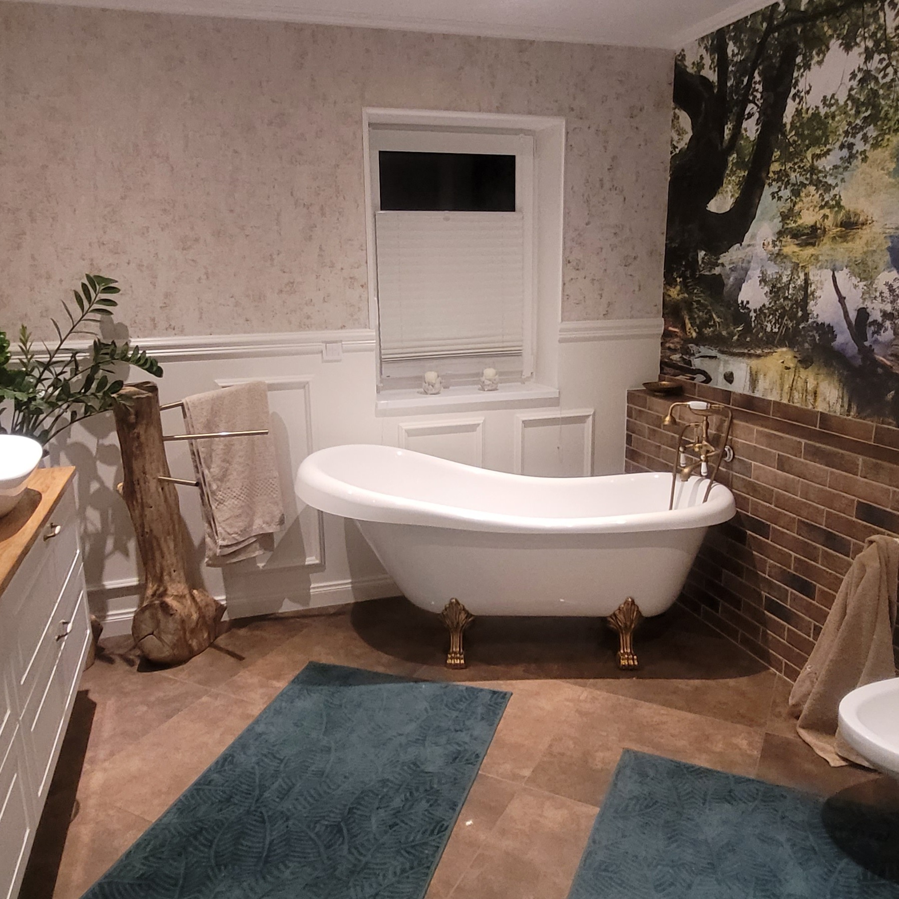 Elegancka łazienka z wolnostojącą wanną na złotych nóżkach, drewnianym wieszakiem na ręczniki z pnia drzewa i fototapetą z motywem leśnym na ścianie.
