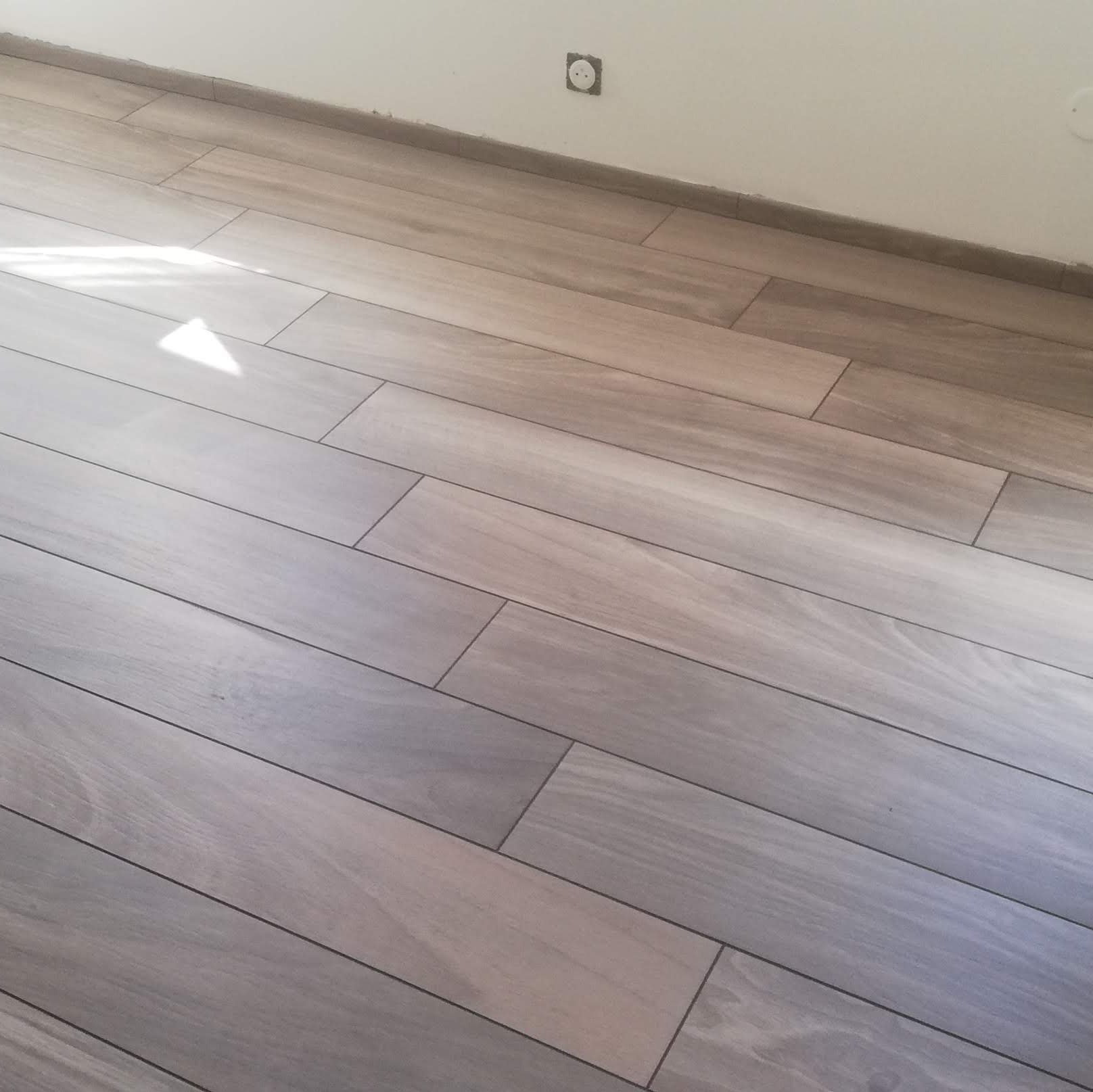 Ułożona pod kątem podłoga z płytek imitujących drewno w odcieniach szarości i beżu, widoczne fugi i listwa przypodłogowa, fragment ściany z gniazdkiem elektrycznym.