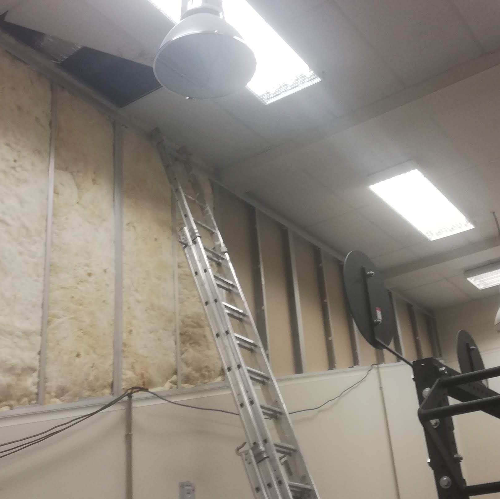 Montaż izolacji z wełny mineralnej w ścianie szkieletowej z widoczną drabiną aluminiową i elementami konstrukcji sufitu podwieszanego w pomieszczeniu biurowym.