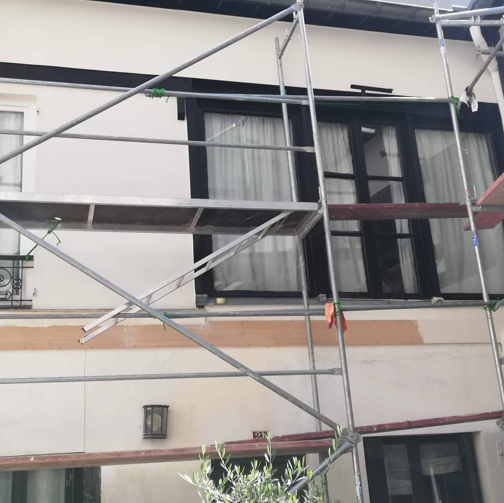 Elewacja budynku w trakcie malowania, widoczne rusztowanie, świeżo pomalowane fragmenty ściany, czarne ramy okienne.