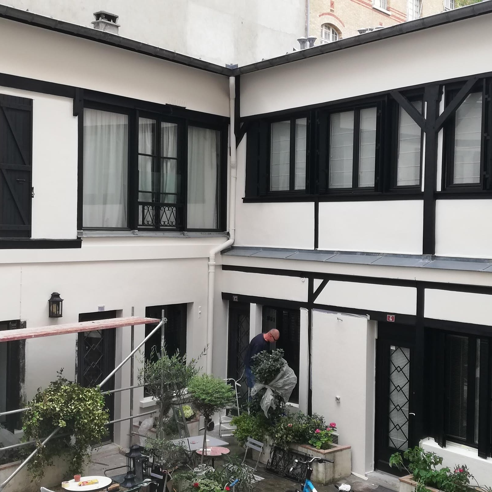 Wewnętrzny dziedziniec budynku po odświeżeniu elewacji, widoczne rusztowanie, rośliny doniczkowe i mężczyzna przenoszący roślinę, białe ściany z czarnymi elementami dekoracyjnymi.