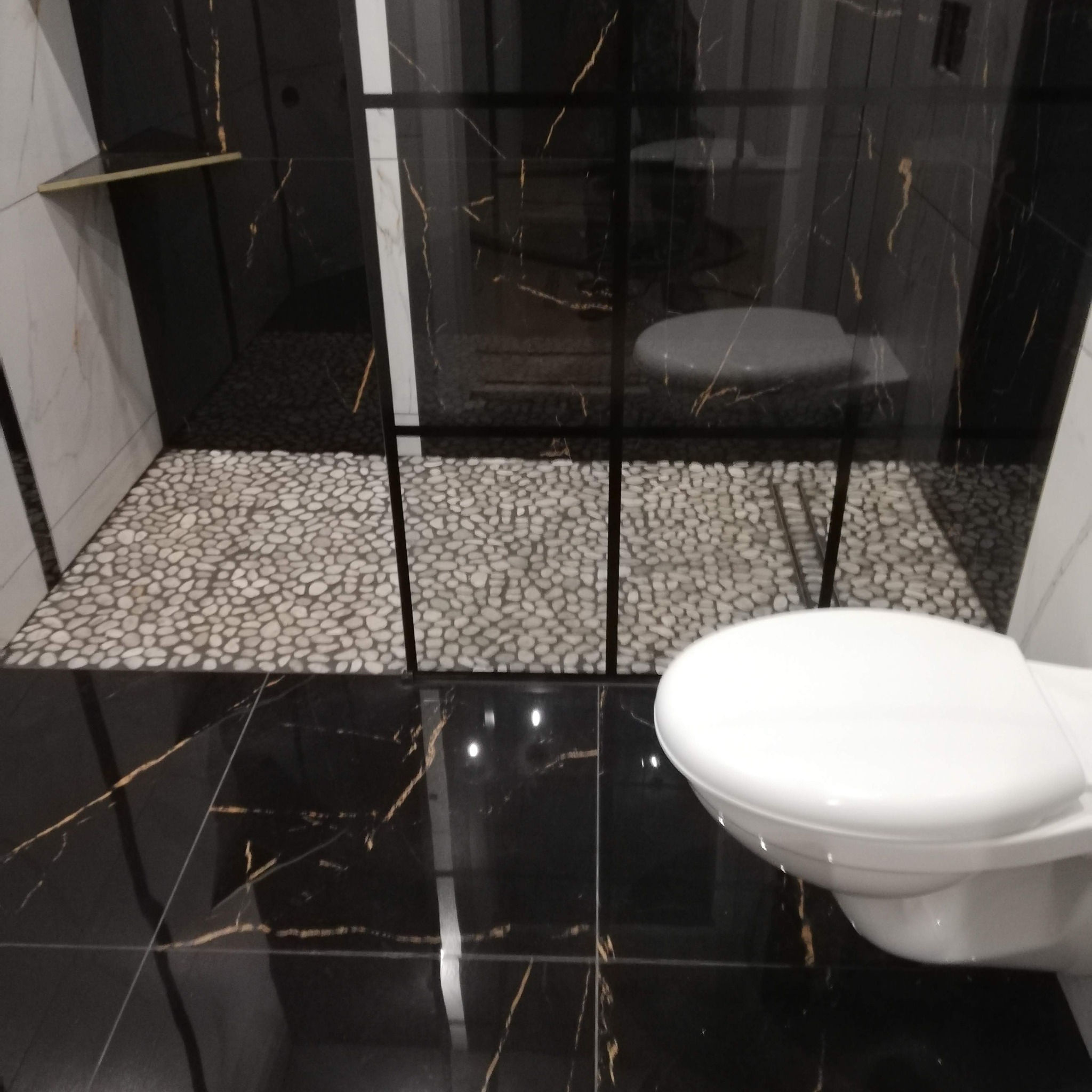 Nowoczesna łazienka z czarnymi marmurowymi płytkami z złotymi żyłkami na podłodze i ścianach, kabina prysznicowa wyłożona białymi kamieniami, widoczny fragment toalety.