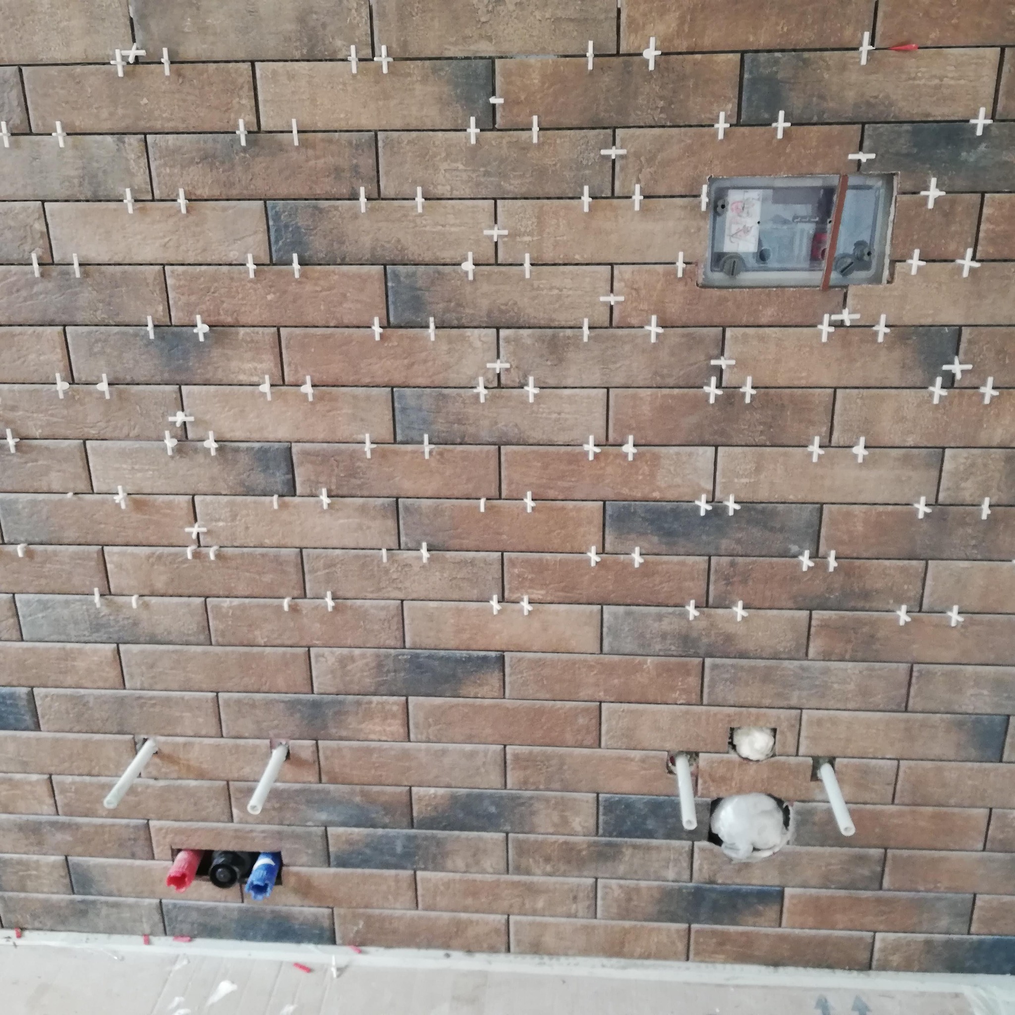 Ściana z brązowo-szarych płytek imitujących cegłę, w trakcie układania, z widocznymi krzyżykami dystansowymi, otworami na instalacje wodne i puszkę elektryczną.