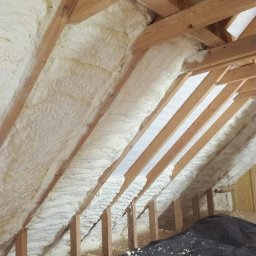 Tynki Maszynowo - Gipsowe Paweł Okoński - Poddasze z drewnianą konstrukcją pokryte pianą izolacyjną, zabezpieczone folią ochronną na podłodze, widoczne białe fragmenty piany na czarnej folii.