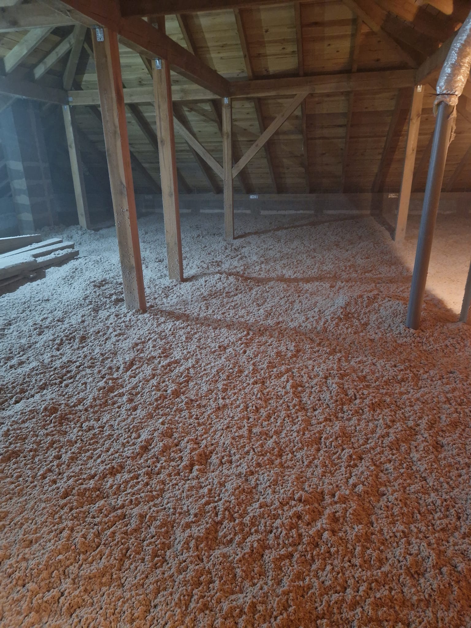 Poddasze z drewnianą konstrukcją pokryte warstwą izolacji z wełny mineralnej, widoczne drewniane słupy konstrukcyjne i fragment instalacji wentylacyjnej.