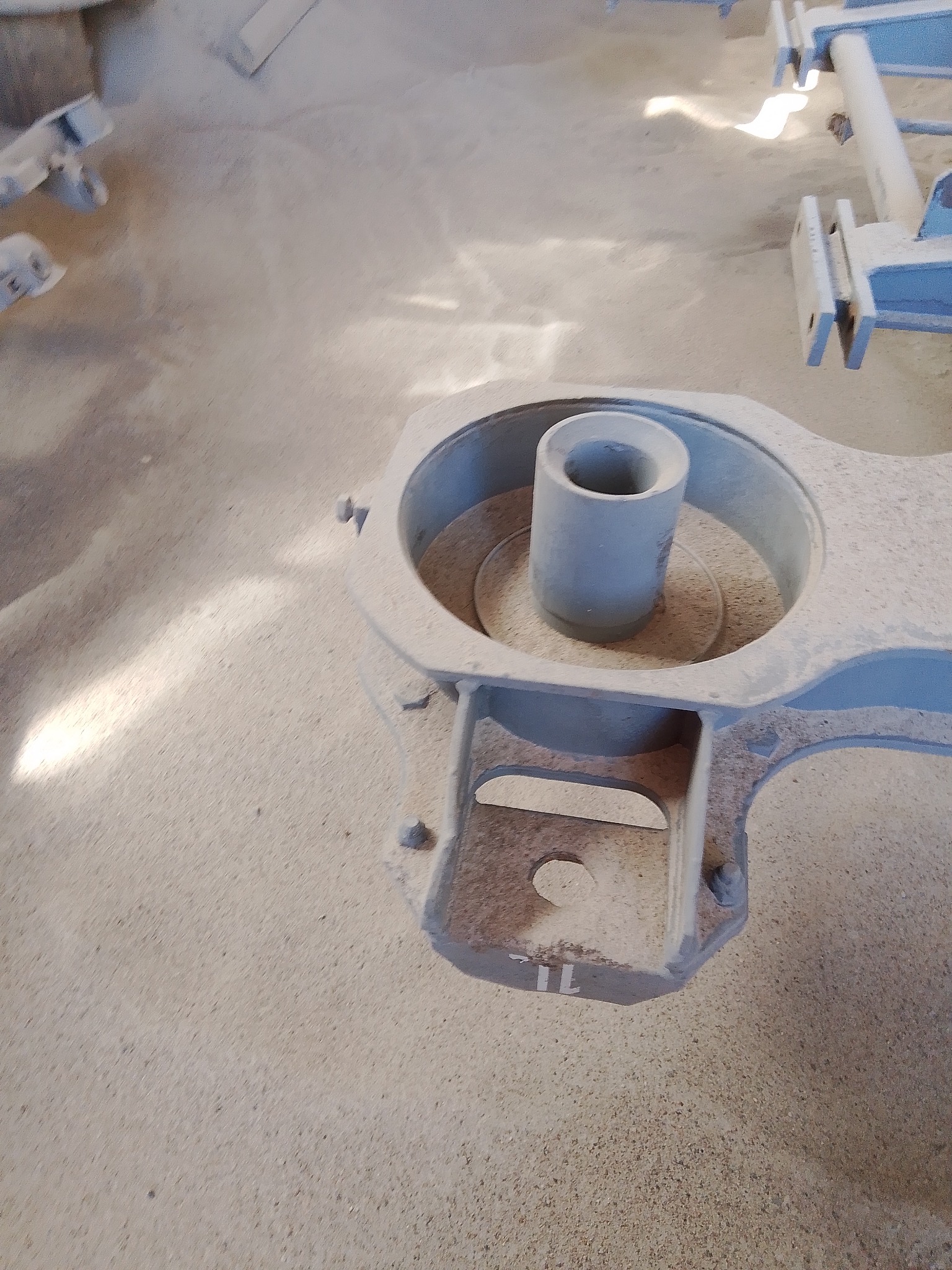 Szara, odlewana forma z cylindrycznym otworem i numerem '1L' na podstawie, otoczona piaskiem odlewniczym.