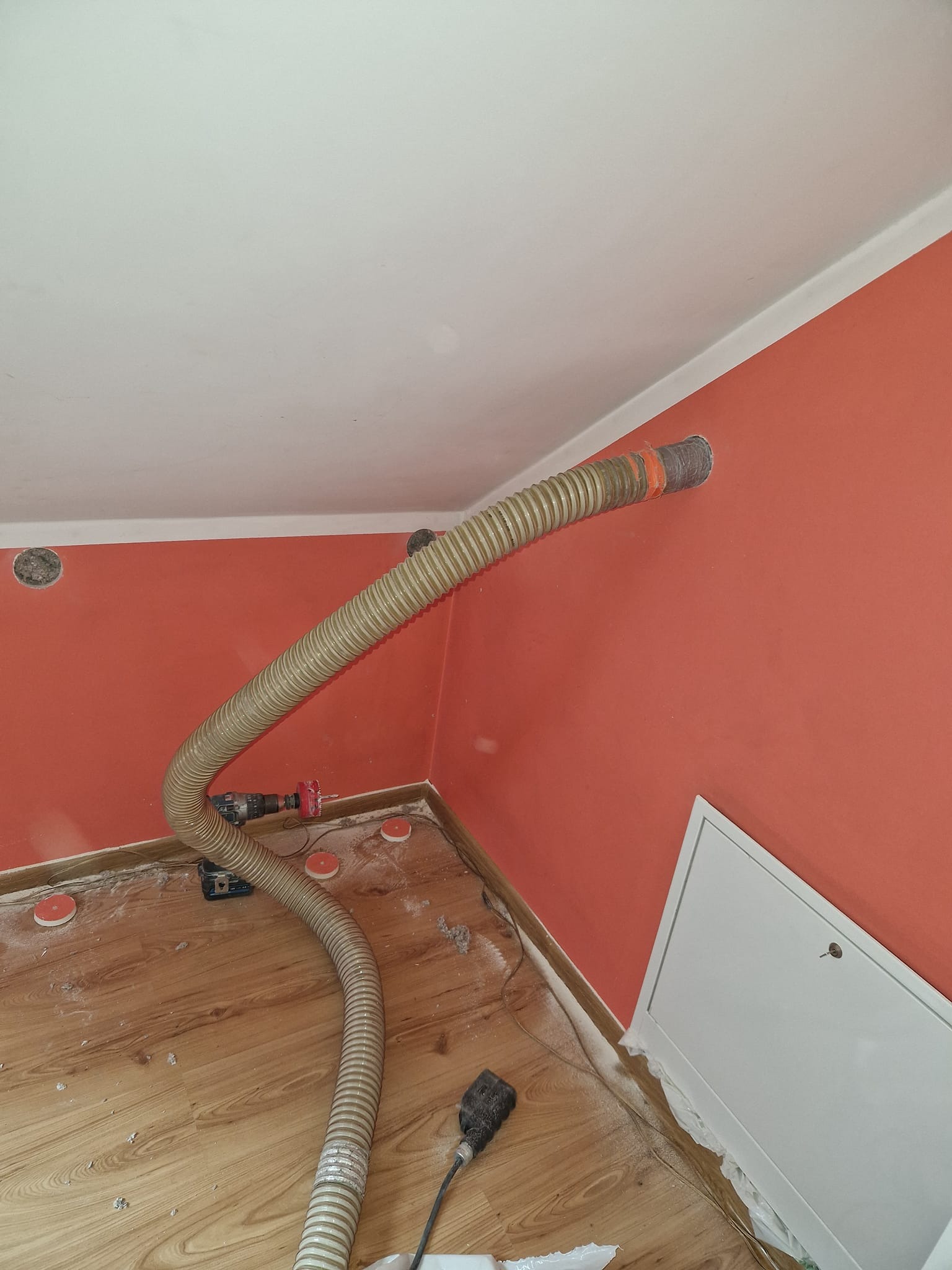 Wąż do wdmuchiwania materiału izolacyjnego w otwór w ścianie, widoczny wiertarko-wkrętak akumulatorowy, zabezpieczone otwory w podłodze, ślady materiału izolacyjnego na podłodze i ścianie.