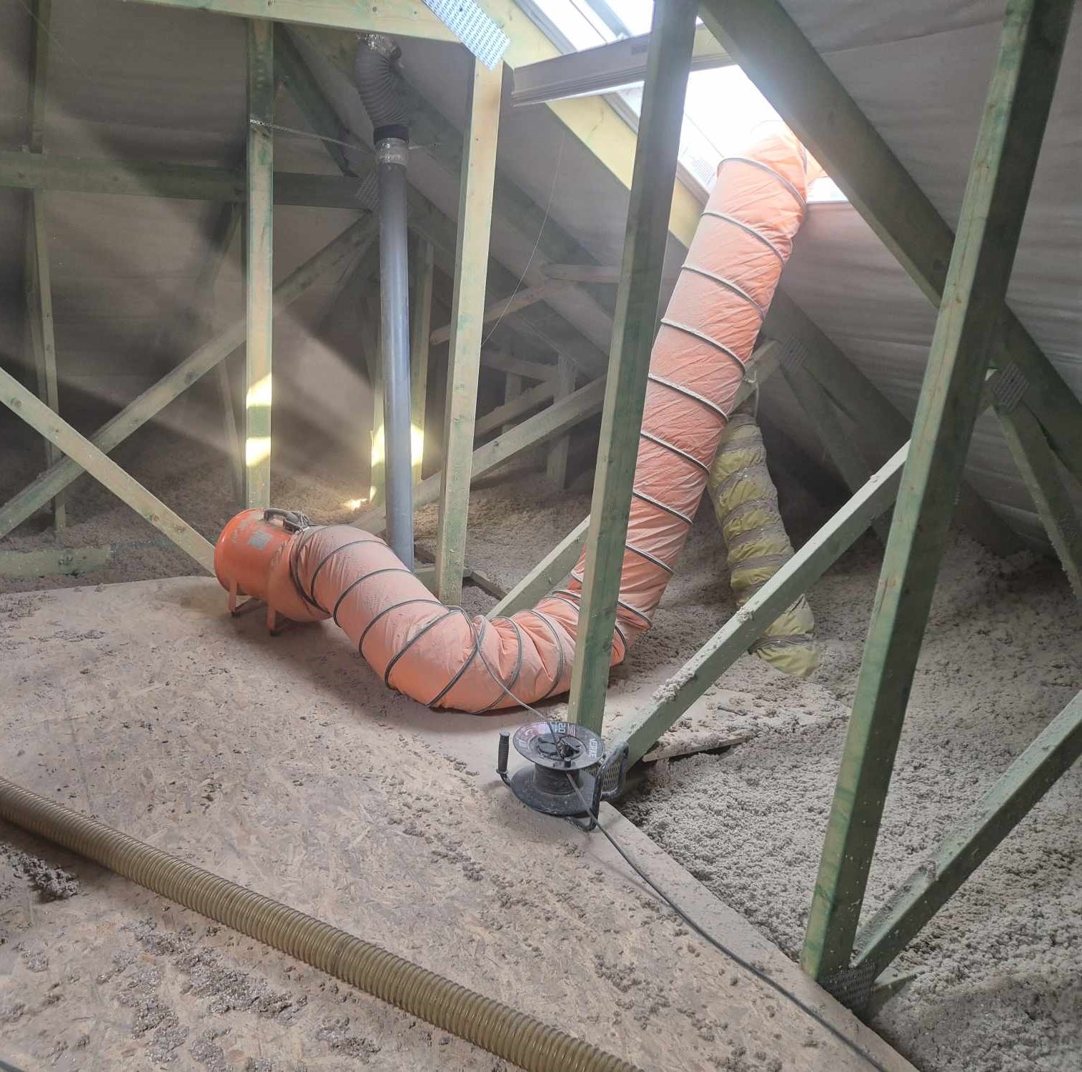 Poddasze w trakcie ocieplania: widoczna warstwa izolacji, pomarańczowy przewód wentylacyjny i drewniana konstrukcja dachu z oknem dachowym w tle.