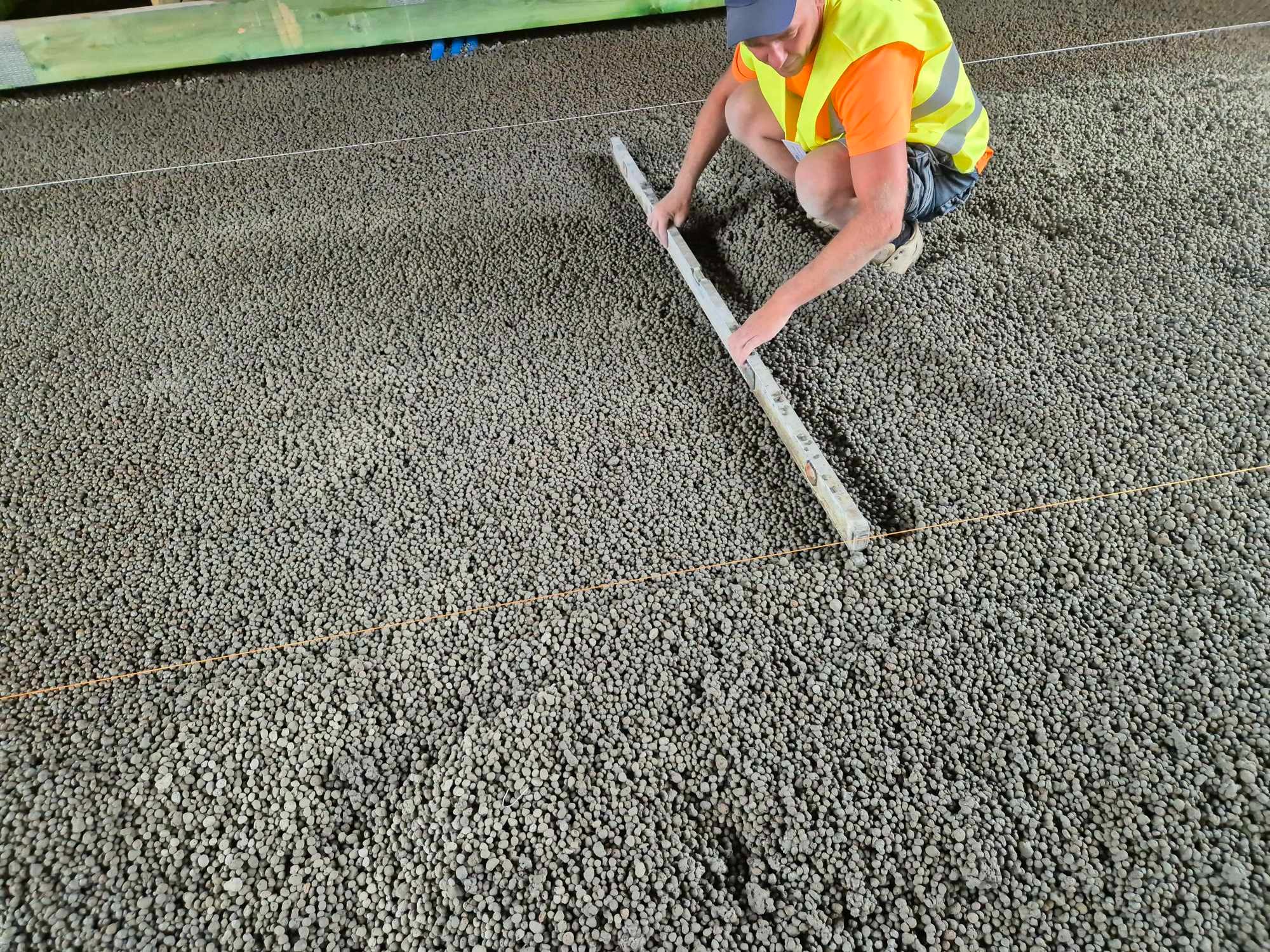 Pracownik w kamizelce odblaskowej wyrównuje podsypkę z granulatu styropianowego za pomocą poziomicy, widoczne sznurki wyznaczające poziom.