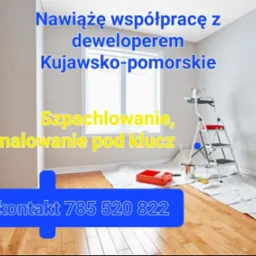 Pokój w trakcie prac wykończeniowych z widoczną drabiną malarską, kuwetą z farbą oraz zabezpieczoną podłogą. Na ścianie widnieje napis informujący o współpracy z deweloperem w Kujawsko-pomorskim...