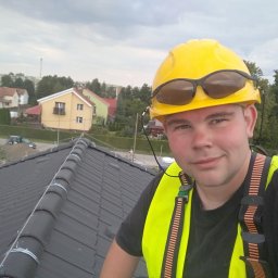 ACDC ALFA Beniamin Korniszuk - Młody dekarz w żółtym kasku i kamizelce odblaskowej, z okularami przeciwsłonecznymi na kasku, na tle nowo położonego, ciemnoszarego dachu z ceramicznej dachówki i zabudowy miejskiej w tle.
