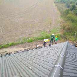 ACDC ALFA Beniamin Korniszuk - Montaż fotowoltaiki na dachu z szarej blachodachówki, widoczne panele montażowe, rura osłonowa i trzech pracowników w kaskach i kamizelkach odblaskowych przygotowujących teren obok dachu.