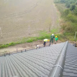 Montaż fotowoltaiki na dachu z szarej blachodachówki, widoczne panele montażowe, rura osłonowa i trzech pracowników w kaskach i kamizelkach odblaskowych przygotowujących teren obok dachu.
