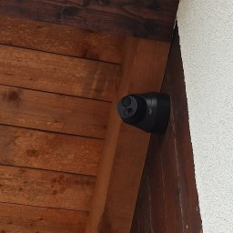 ACDC ALFA Beniamin Korniszuk - Czarna kamera monitoringu marki UNIV zamontowana pod drewnianym okapem, widoczne dwie soczewki i fragment białego tynku ściany.