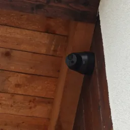 Czarna kamera monitoringu marki UNIV zamontowana pod drewnianym okapem, widoczne dwie soczewki i fragment białego tynku ściany.