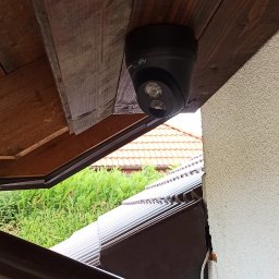 ACDC ALFA Beniamin Korniszuk - Czarna kamera monitoringu marki Uniview zamontowana pod drewnianym okapem, widok z dołu na obiektyw, z fragmentem elewacji budynku i dachu w tle.