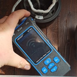 ACDC ALFA Beniamin Korniszuk - Miernik optyczny do kabli NOYAFA NF-8508, z ekranem wskazującym tryb cyfrowy, trzymany w dłoni na tle drewnianej ściany i fragmentu drabiny, obok czarnej puszki instalacyjnej z podłączonym przewodem.