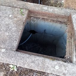 ACDC ALFA Beniamin Korniszuk - Szary przewód w wykopanym rowie ziemnym, wchodzący do betonowej studzienki z otwartym wiekiem, widok z góry, częściowo widoczne buty osoby robiącej zdjęcie.