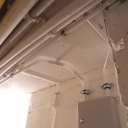 ACDC ALFA Beniamin Korniszuk - Białe rury instalacyjne prowadzone pod sufitem wzdłuż ściany z zamontowaną szarą skrzynką elektryczną z napisem 'Touetic' poniżej zamka.