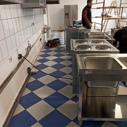 ACDC ALFA Beniamin Korniszuk - Wnętrze kuchni komercyjnej w trakcie instalacji: widoczne stalowe blaty z wmontowanymi płytami grzewczymi, zlewozmywaki, podłączenia elektryczne wzdłuż ściany i pracownicy na rusztowaniu w tle.