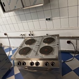 ACDC ALFA Beniamin Korniszuk - Dwie czteropalnikowe, elektryczne płyty grzewcze ze stali nierdzewnej marki Hendi, ustawione w kuchni z białymi kafelkami i niebiesko-białą podłogą w kratkę; widoczne gniazdka elektryczne i rury.