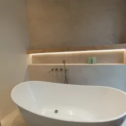 Nowoczesna, wolnostojąca wanna owalna w łazience wykończonej płytkami imitującymi kamień, z podświetlaną półką nad wanną i minimalistyczną armaturą w kolorze starego złota.