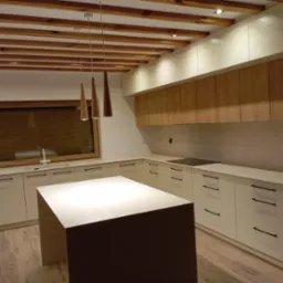 Nowoczesna, jasna kuchnia z drewnianymi akcentami: wyspa, szafki wiszące z drewnianymi frontami, drewniane belki na suficie i trzy wiszące lampy o stożkowym kształcie.