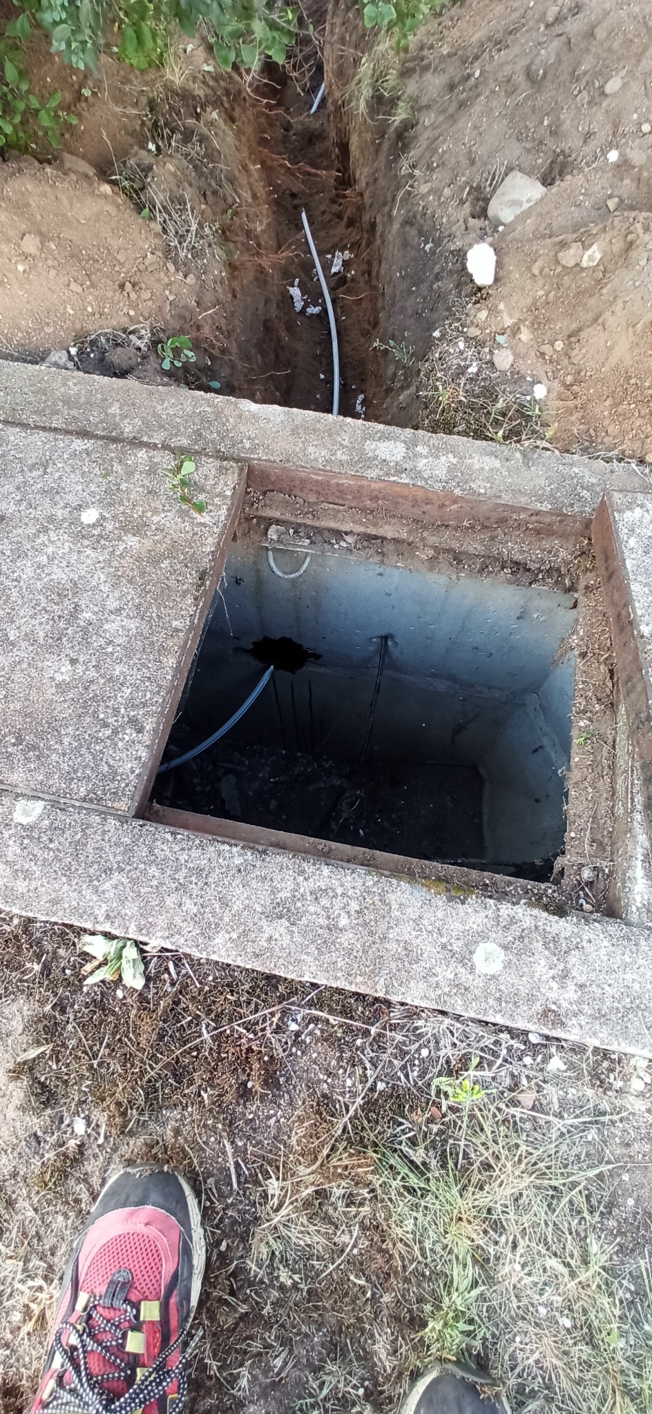 Szary przewód w wykopanym rowie ziemnym, wchodzący do betonowej studzienki z otwartym wiekiem, widok z góry, częściowo widoczne buty osoby robiącej zdjęcie.