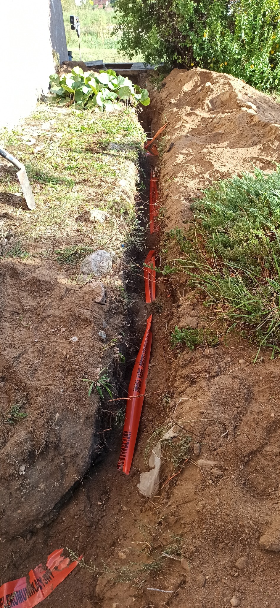 Wkopane pomarańczowe rury w wykopie ziemnym, zabezpieczone taśmą ostrzegawczą 'ELEKTROKOMUNIKACYJNY', widok z góry na świeżo przekopaną ziemię i fragment roślinności.