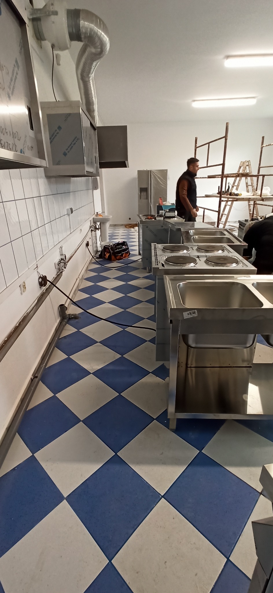 Wnętrze kuchni komercyjnej w trakcie instalacji: widoczne stalowe blaty z wmontowanymi płytami grzewczymi, zlewozmywaki, podłączenia elektryczne wzdłuż ściany i pracownicy na rusztowaniu w tle.