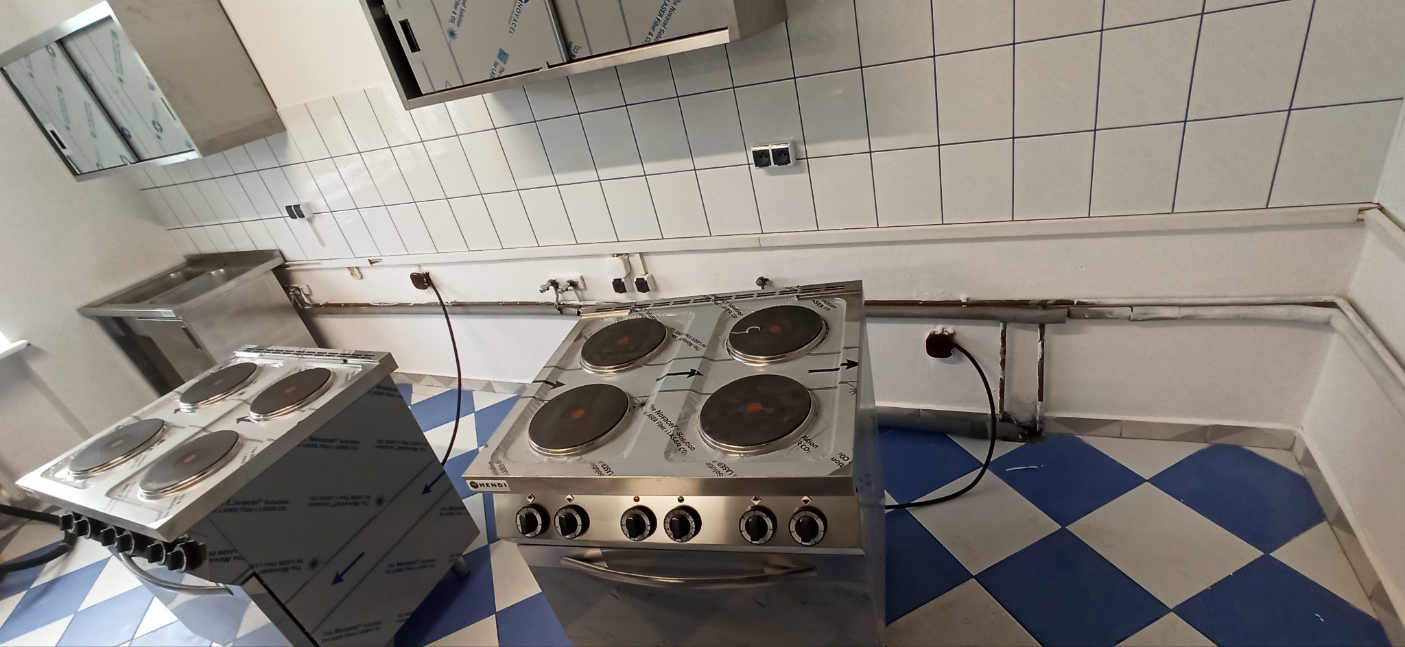 Dwie czteropalnikowe, elektryczne płyty grzewcze ze stali nierdzewnej marki Hendi, ustawione w kuchni z białymi kafelkami i niebiesko-białą podłogą w kratkę; widoczne gniazdka elektryczne i rury.