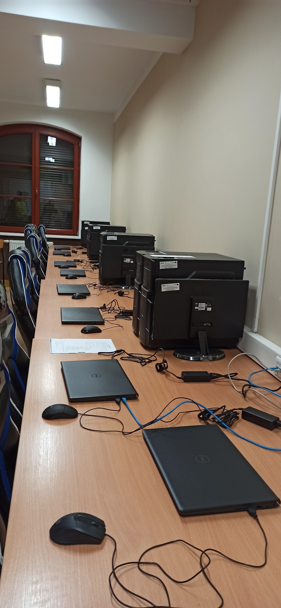 Sala komputerowa z rzędem biurek, na których stoją laptopy Dell, myszki, monitory LG i komputery stacjonarne, widoczne kable zasilające i sieciowe, oświetlenie sufitowe oraz okno z roletami.