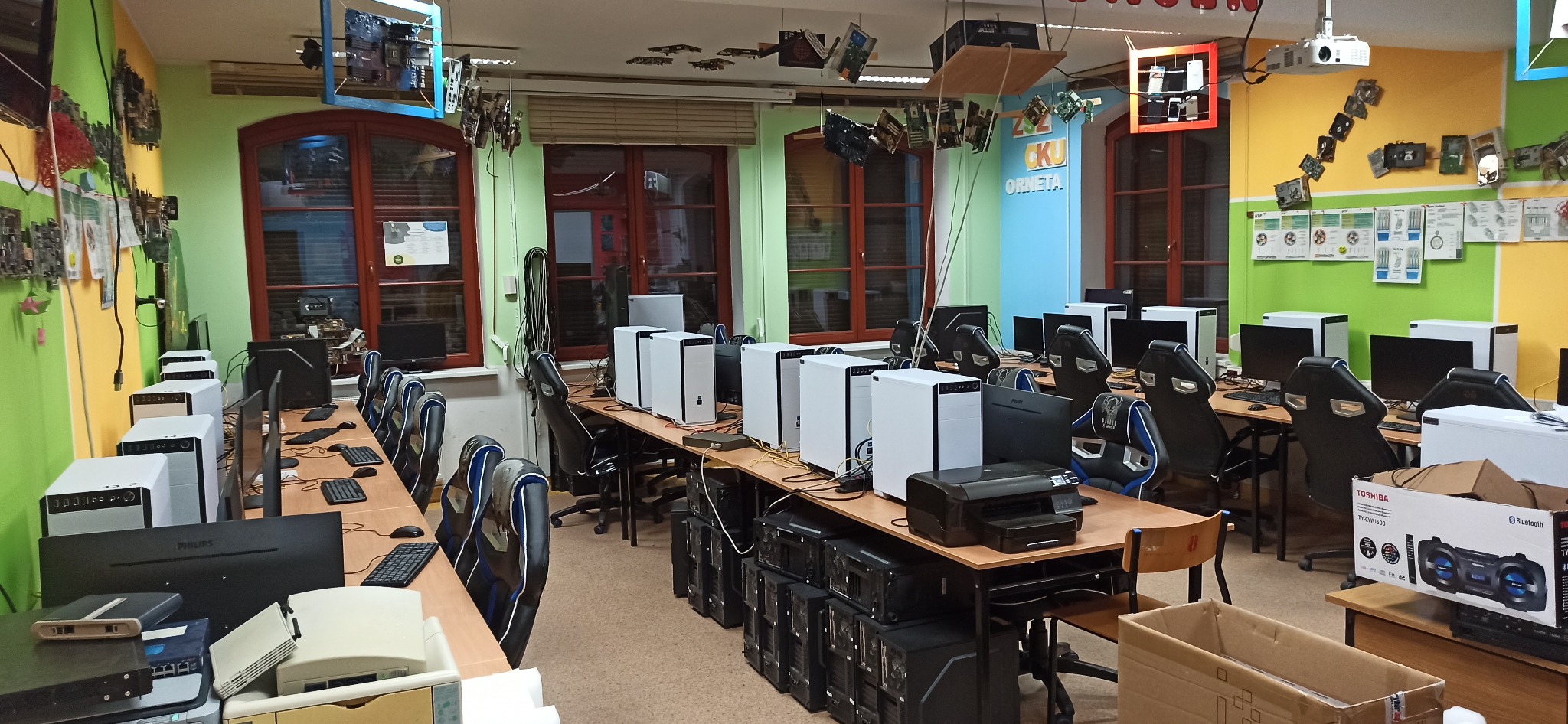 Sala komputerowa z rzędami biurek, na których stoją białe obudowy komputerów, monitory i klawiatury. Ściany zdobią ramki z podzespołami komputerowymi i schematami. Projektor zamontowany pod sufitem.
