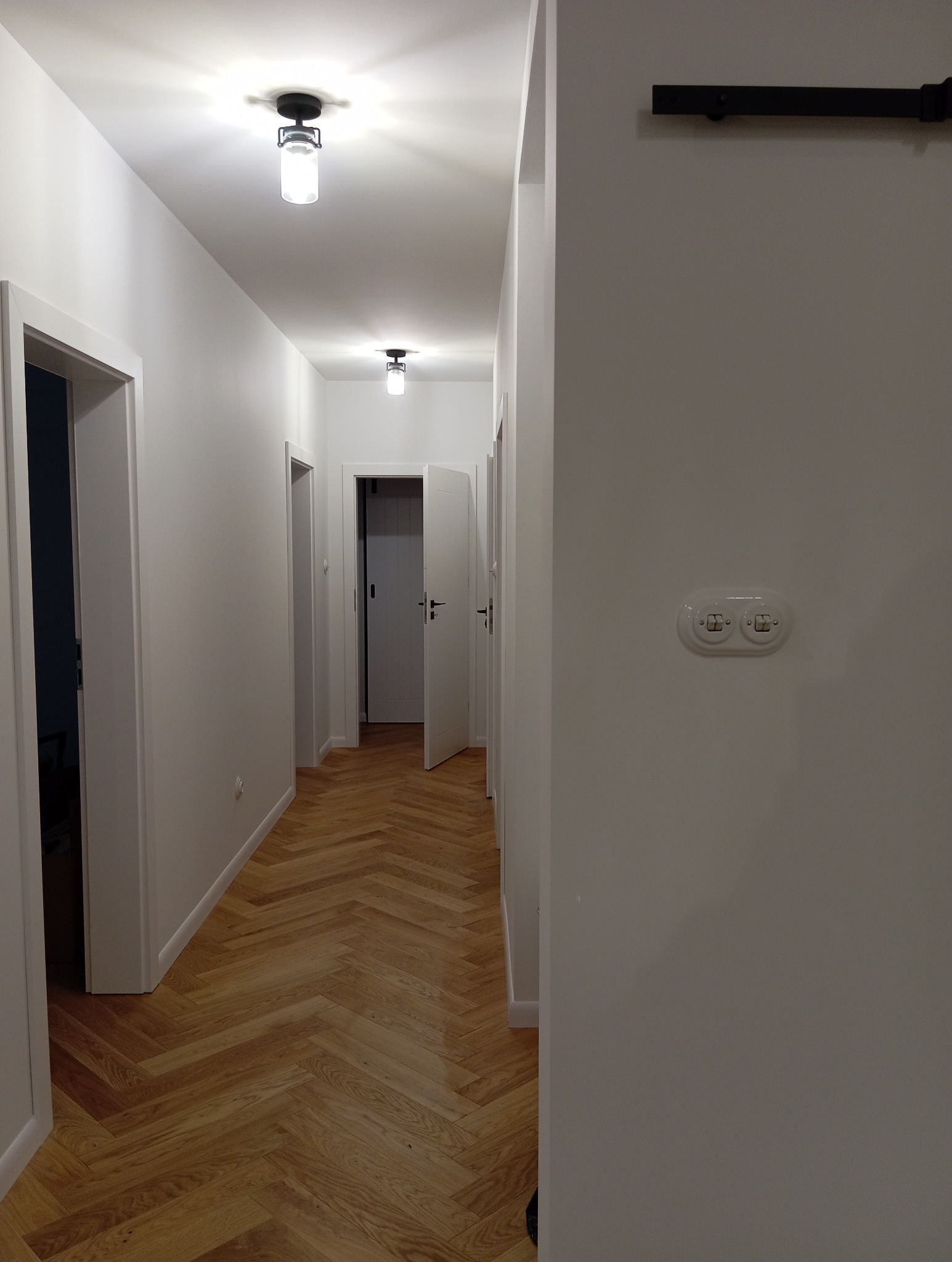 Nowoczesny korytarz z parkietem w jodełkę, białymi ścianami i minimalistycznym oświetleniem sufitowym z cylindrycznymi kloszami. Na ścianie widoczny włącznik światła w stylu retro.