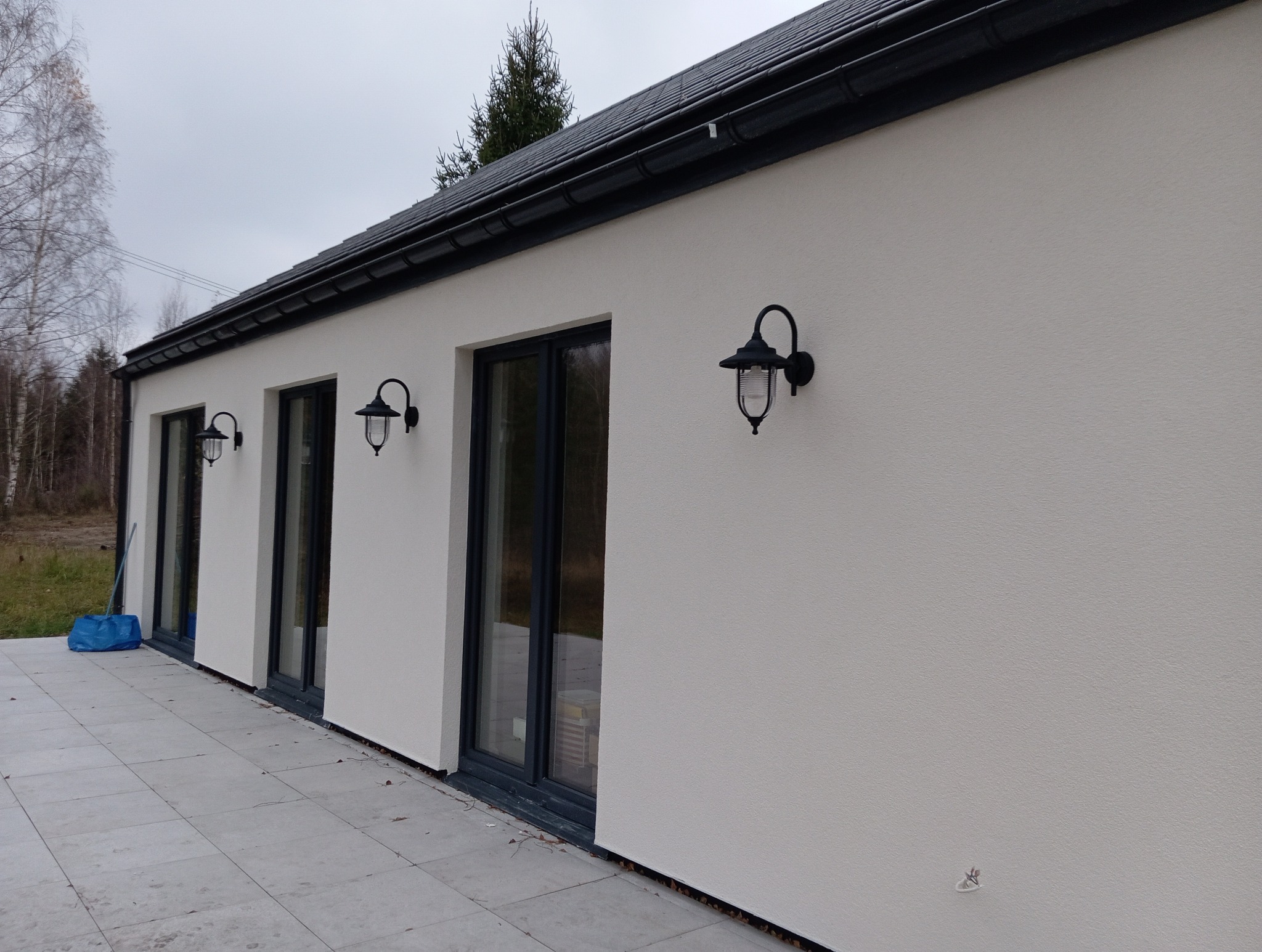 Elewacja domu jednorodzinnego z trzema oknami i zamontowanymi czarnymi lampami zewnętrznymi w stylu retro, widoczny fragment dachu z czarną dachówką i szarym niebem.