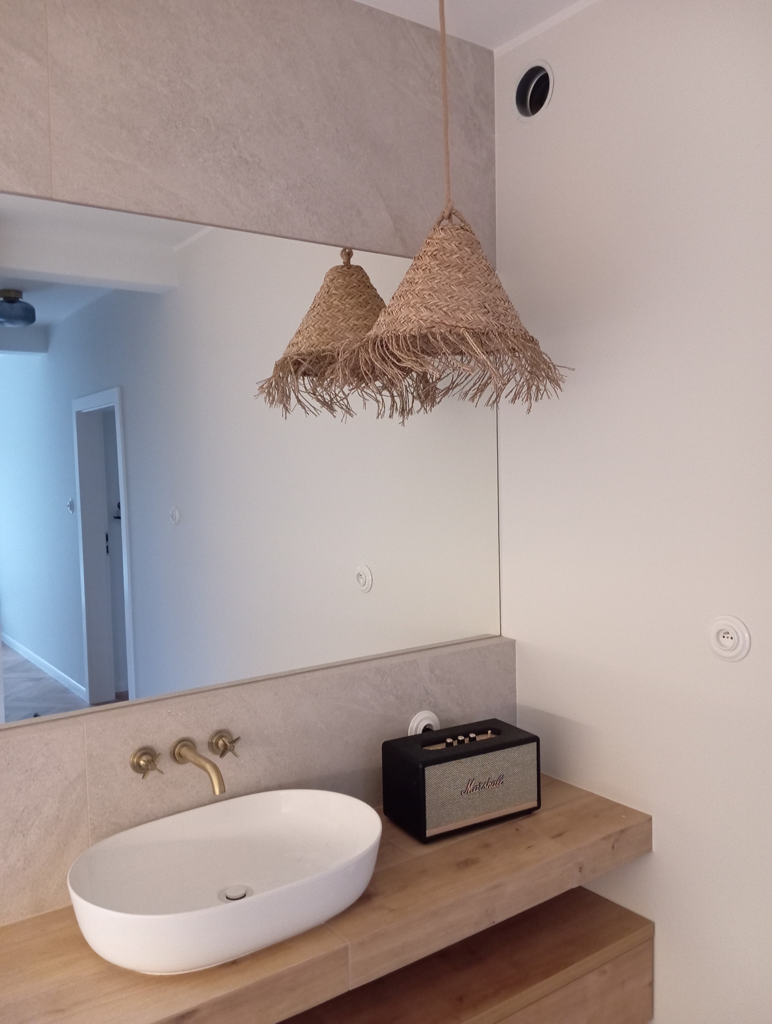 Nowoczesna łazienka z owalną umywalką nablatową, złotą baterią ścienną i lampami wiszącymi z naturalnego włókna, odbijającymi się w lustrze.