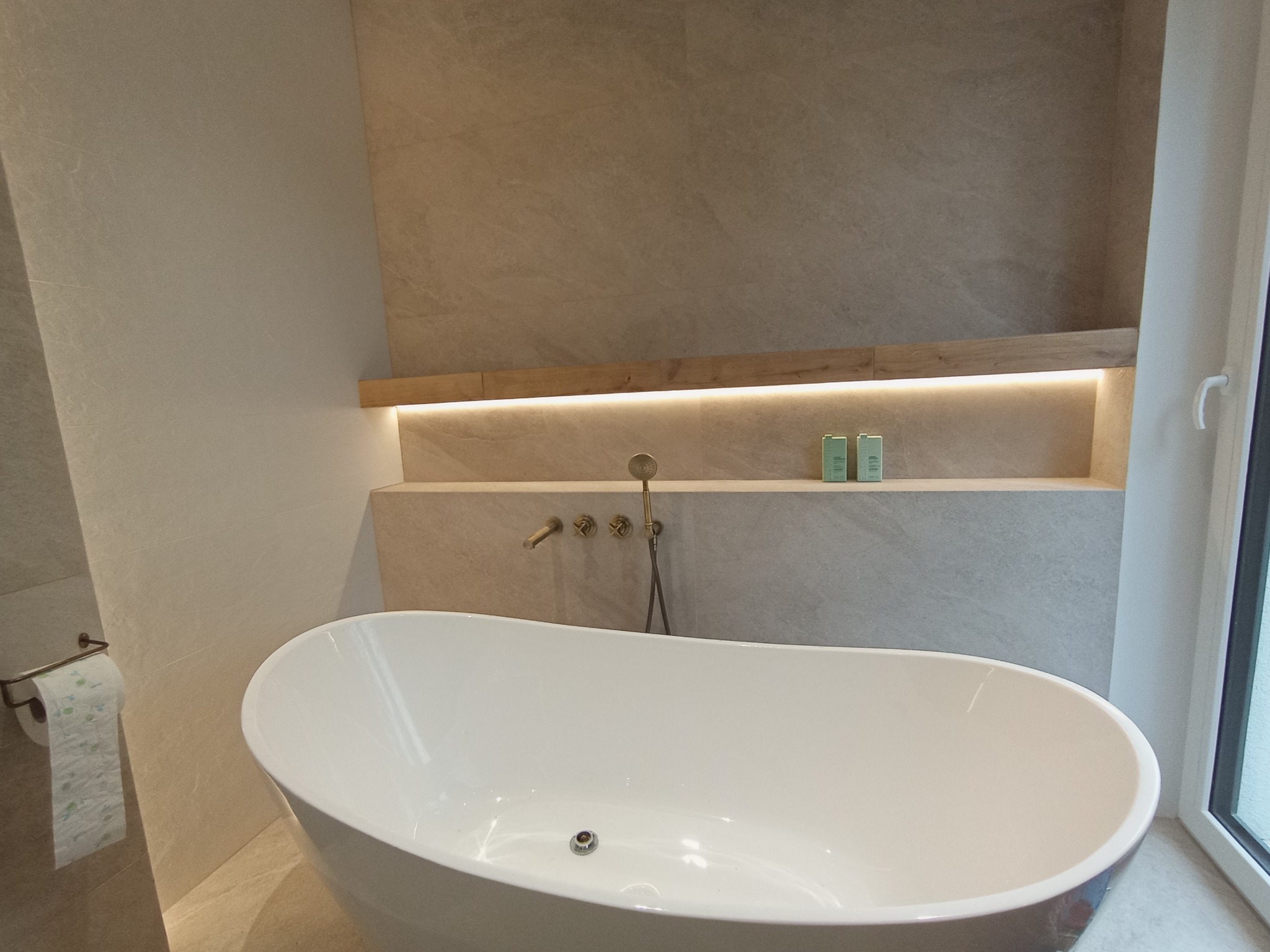 Nowoczesna, wolnostojąca wanna owalna w łazience wykończonej płytkami imitującymi kamień, z podświetlaną półką nad wanną i minimalistyczną armaturą w kolorze starego złota.