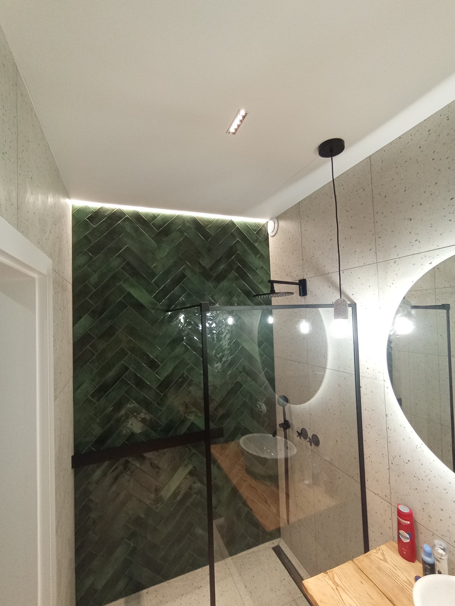 Nowoczesna łazienka w Lidzbarku Warmińskim z zielonymi płytkami ułożonymi w jodełkę, kabina prysznicowa z czarnymi ramami, okrągłe lustro z podświetleniem i designerska lampa wisząca.