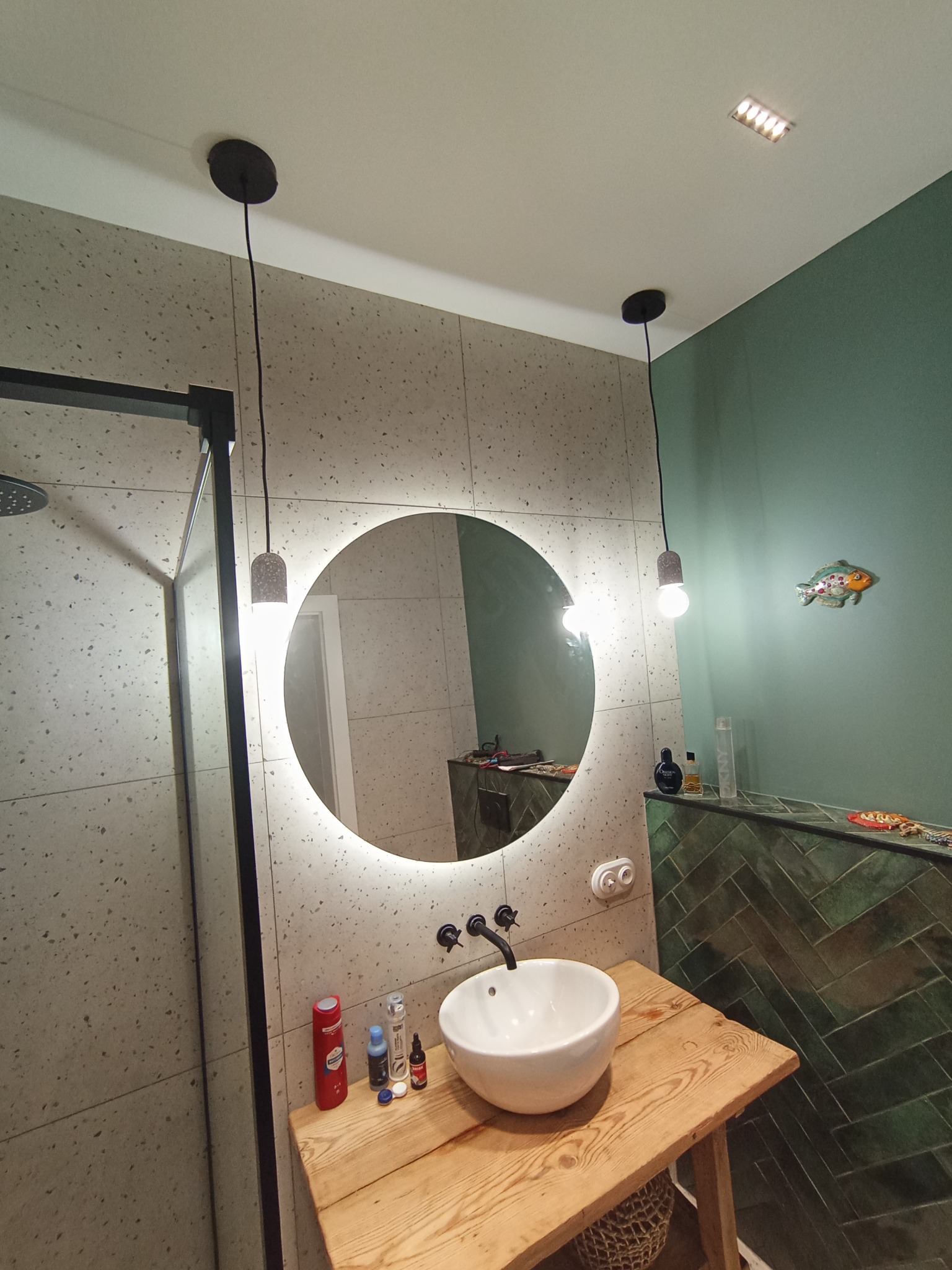 Nowoczesna łazienka z okrągłym lustrem podświetlanym LED, umywalką nablatową na drewnianym blacie i wiszącymi lampami z betonowymi kloszami. Ściany wyłożone płytkami imitującymi lastryko...