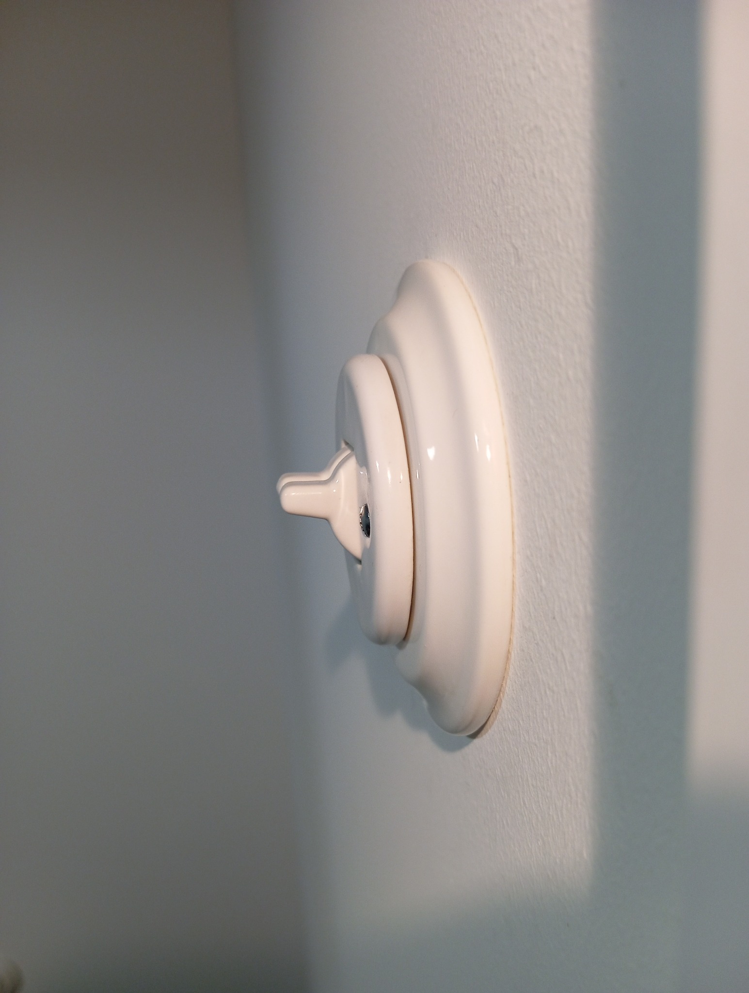 Biały, porcelanowy włącznik światła na białej, teksturowanej ścianie, oświetlony bocznym światłem, rzucającym cień.